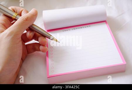 Linke Hand hält einen Kugelschreiber und schreibt auf einem 100 Prozent wichtigen rosa Notizblock Stockfoto
