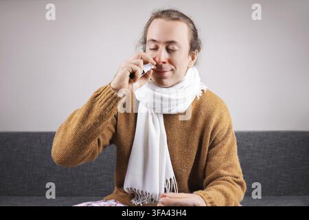 Kranker Mann mit roter Nase verwendet Nasenspray zur Behandlung von Schnupfen und Verstopfung, während er zu Hause auf dem Sofa sitzt Stockfoto