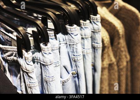 Nahaufnahme von hellblauen Denimjeans und Hosen, die auf schwarzen Kleiderbügeln neben einem beigen Strickpullover in einem Bekleidungsgeschäft hängen Stockfoto