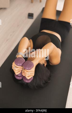 Frau hält Hanteln über dem Kopf, während sie auf einer Yogamatte liegt, und trainiert drinnen Stockfoto