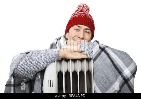 Glücklicher junger Mann mit Wintermütze und Decke, der sich in der Nähe eines elektrischen Heizgeräts aufwärmt. Vor weißem Hintergrund Stockfoto