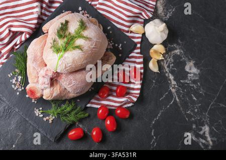 Ansicht von oben eines ganzen gefrorenen rohen Huhns auf einem schwarzen Schieferteller mit Kirschtomaten, Knoblauch und Dill. Konzept der Lebensmittellagerung, gefrorenes Geflügel an Stockfoto