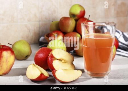 Ein Glas frischer Apfelsaft auf einer Marmorküchenarbeitsplatte, begleitet von ganzen und geschnittenen roten Äpfeln in einem Weidenkorb. Stockfoto