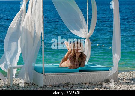 Frauenstrandhütte, eine ruhige Frau entspannt sich in einer weißen Strandhütte am funkelnden blauen Ozean. Stockfoto