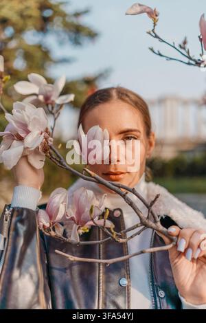 Magnolia-Frau im Frühling hält einen blühenden Ast vor ihrem Gesicht in einem sonnigen Park während des Sonnenuntergangs Stockfoto
