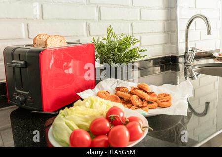 Roter Toaster mit Brot neben Gemüse und gegartem paniertem Fisch in der Küche Stockfoto
