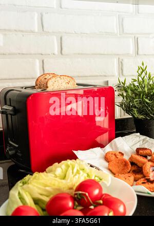 Roter Toaster mit Brot neben Gemüse und gegartem paniertem Fisch in der Küche Stockfoto