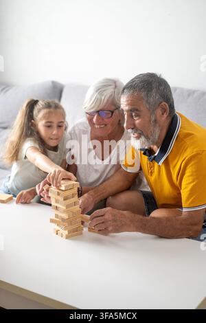 Zufriedene Großeltern spielen Holzklotzturm-Spiel mit ihren Enkelinnen zu Hause Stockfoto