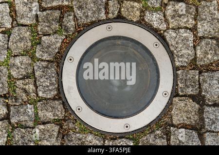 Rundes bodengrabenes Lichtfixture, eingebettet in strukturierten grauen Granitkobelsteinbelag, urbanes Außenbeleuchtungskonzept Stockfoto