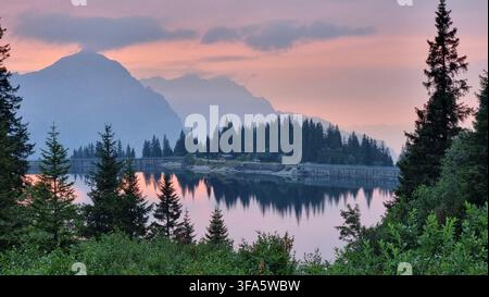 Malereischer Bergsee, umgeben von immergrünem Wald und dramatischen Bergen, die sich im ruhigen Wasser bei Dämmerung spiegeln. Stockfoto