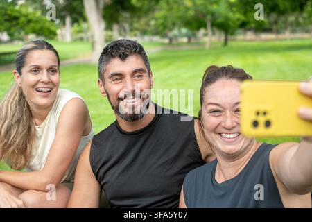 Drei lächelnde Freunde machen ein Gruppen-Selfie mit einem Smartphone, während sie nach dem gemeinsamen Sport in einem grünen Park entspannen. Stockfoto