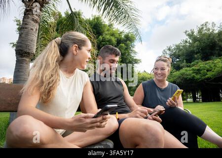 Gruppe von Freunden, die auf einer Parkbank sitzen, Smartphones benutzen und miteinander reden, während sie nach sportlicher Betätigung im Freien eine Pause machen. Stockfoto