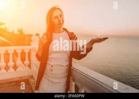 Sonnenuntergangsfrau Balkon steht auf einer Terrasse mit Blick auf das Meer während der goldenen Stunde und hält die Sonne in ihrer Hand Stockfoto
