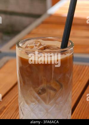 Nahaufnahme eines erfrischenden Eiskaffee-Latte mit Milch und Eiswürfeln in einem hohen Glas mit einem Strohhalm auf einem Holztisch. Stockfoto