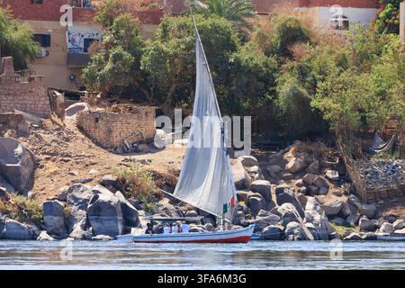 Touristen reisen in einem traditionellen Felukka-Segelboot auf dem Nil in der Nähe von Assuan, Ägypten, mit dem felsigen Flussufer und dem Dorf am Ufer im B Stockfoto