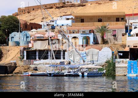 Nubische Gästehäuser und traditionelle Häuser am Ufer des Nils in der Nähe von Assuan, Ägypten, mit farbenfrohen Gebäuden, lokalen Booten und festgemachten Felukkas Stockfoto