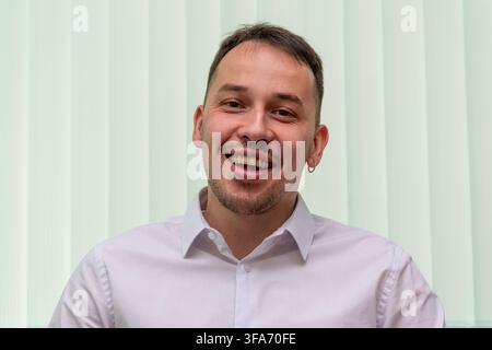 Ein lächelnder junger Mann in einem weißen Hemd schaut in die Kamera vor einem hell gestreiften Hintergrund. Stockfoto