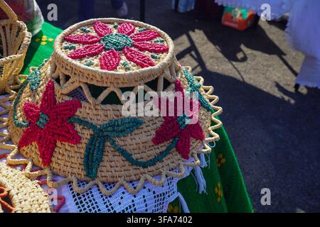 Handgefertigter geflochtener Strohkorb mit bestickten roten Blumenmustern. Traditionelles griechisches Handwerk und Volkskunstprodukt, das auf einer grünen Tischdecke ausgestellt ist. Stockfoto