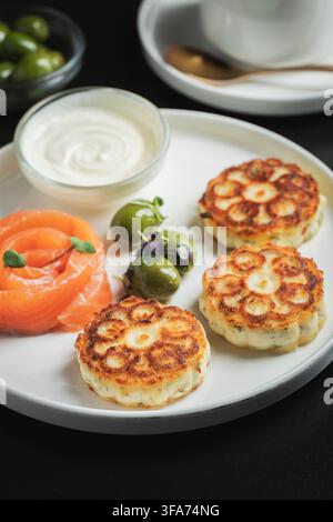 Herzhafte Syrniki mit Räucherlachs, Oliven und Sauerrahm auf weißer Platte Stockfoto