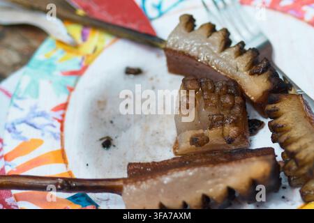 Nahaufnahme von gebratenem Schweinefett und Bacon-Scheiben, die auf Holzstäbchen serviert werden, über einem gemusterten Pappteller mit einer Gabel in der Nähe. Stockfoto