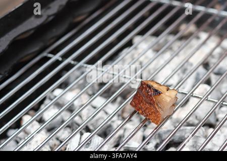 Eine Nahaufnahme eines einzelnen kleinen Stücks herzhaftem Speck, das auf einem Metall-Grillrost über glühenden Holzkohleembern brät. Stockfoto