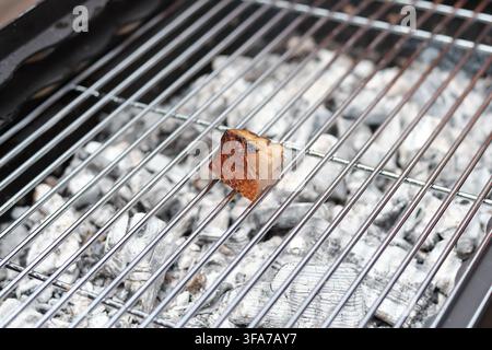 Eine Nahaufnahme eines einzelnen kleinen Stücks herzhaften Specks, der auf einem glänzenden Metallgrillrost über heißen weißen Holzkohleaschen röstet. Stockfoto