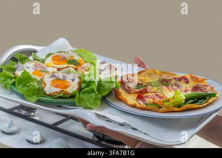Eine Person hält ein weißes Tablett mit zwei Tellern Eiern. Ein Teller hat gebratene Eier auf Salat, und der andere hat ein Omelett mit Fleisch und Gemüse. Stockfoto