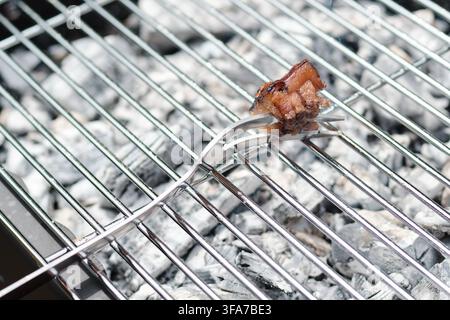 Eine Nahaufnahme eines einzelnen Stücks knusprigem Speck, das auf einer Metallgabel über einem heißen Holzkohlegrillrost mit glühenden Kohlen und Rauch gehalten wird. Stockfoto
