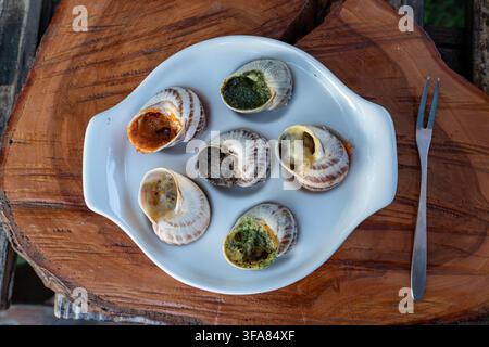 Gebackene gefüllte Mollusken werden mit grünem Kräuterpesto und roter scharfer Tomatensoße in einem speziellen weißen Tablett auf Holzunterlage neben einer Metallgabel präsentiert Stockfoto