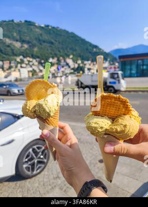 Zwei Hände halten italienische Gelato-Waffelkegel mit einem Hintergrund aus dem Comer See und Bergen in Italien. Stockfoto