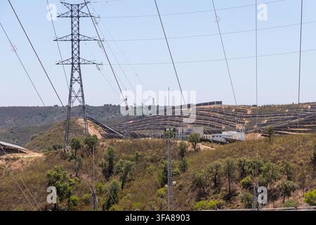 Reihen von Solarpanelen sind auf einem Hügel mit Stromübertragungsmasten und Leitungen im Vordergrund installiert Stockfoto