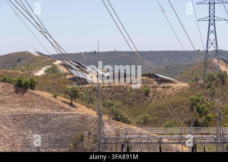 Reihen von Solarpanelen sind in einer sonnengetränkten, trockenen Landschaft installiert, während Hochspannungsleitungen sich über das Bild erstrecken Stockfoto