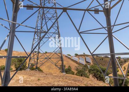 Eine große Metallpylon-Struktur rahmt eine Ansicht von Solarmodulen ein, die auf einem ausgetrockneten, grasbewachsenen Hang mit verstreuten Bäumen und einem hellen Himmel installiert sind Stockfoto