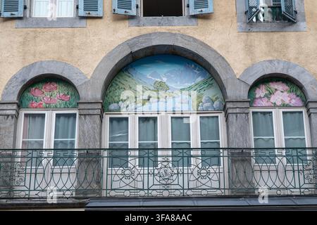 Eine detaillierte architektonische Außenansicht zeigt dekorative gemalte Fresken über drei gewölbten Fenstern an einer historischen europäischen Gebäudefassade. Die c Stockfoto