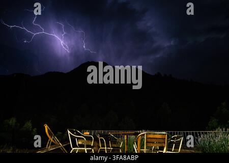 Eine dramatische Nachtszene, die lebhafte Blitze zeigt, die dunkle violette Gewitterwolken über einer silhouettierten Bergkette erhellen. Eine leere Außenterrasse mit Stockfoto