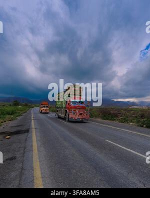 Blick auf zwei traditionell dekorierte pakistanische Lastwagen, die auf einer Straße fahren, mit den majestätischen Sulaiman-Bergen im Hintergrund unter einem dramatischen bewölkten Himmel Stockfoto