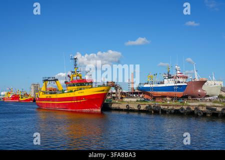 Ventspils, Lettland - 09. August 2026: Bunte Fischerboote, die neben Schiffen unter Reparatur im Industriehafen von Ventspils im Hafen von Lettland liegen Stockfoto