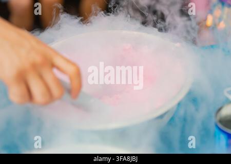 Hand, die mit einer Metallschaufel ein rosa gefrorenes Gelato-Dessert mit flüssigem Stickstoff-Nebel-Effekt in einer Edelstahl-Schale für Eventcatering zubereitet Stockfoto