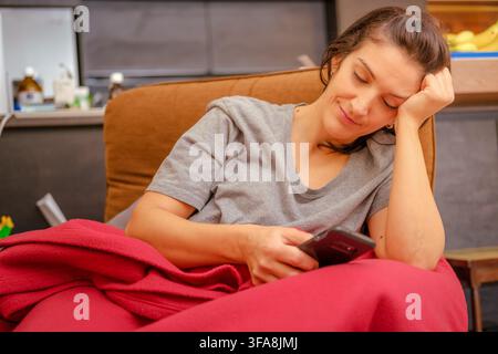 Junge Frau ruht auf dem Sofa unter einer Decke und benutzt ein Mobiltelefon Stockfoto