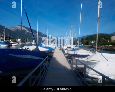 Ein malerisches Yachthafen mit festgemachten Segelbooten und Yachten an einem ruhigen Alplensee, mit Bergen und einem historischen Turm im Hintergrund. Perfekt für Stockfoto