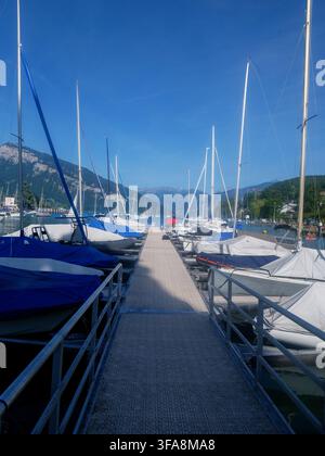 Ein malerisches Yachthafen mit festgemachten Segelbooten und Yachten an einem ruhigen Alplensee, mit Bergen und einem historischen Turm im Hintergrund. Perfekt für Stockfoto