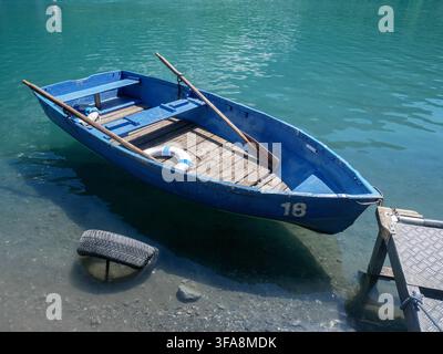 Ein blaues Holzruderboot ruht am Ufer eines türkisfarbenen Alpenssees, umgeben von Bergen, bereit zur Vermietung. Perfekt für Reisen, Freizeit und die Schweiz Stockfoto