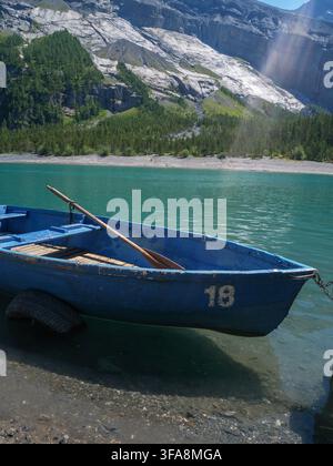 Ein blaues Holzruderboot ruht am Ufer eines türkisfarbenen Alpenssees, umgeben von Bergen, bereit zur Vermietung. Perfekt für Reisen, Freizeit und die Schweiz Stockfoto