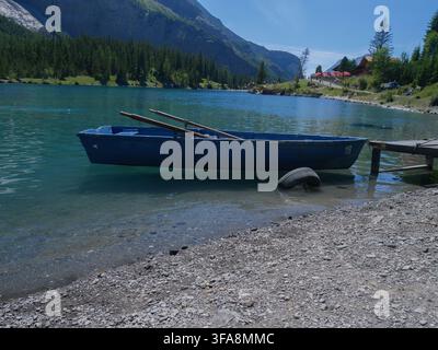 Ein blaues Holzruderboot ruht am Ufer eines türkisfarbenen Alpenssees, umgeben von Bergen, bereit zur Vermietung. Perfekt für Reisen, Freizeit und die Schweiz Stockfoto