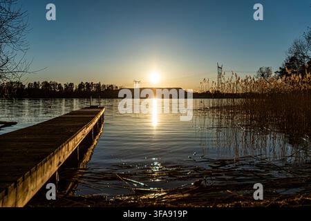 Ein hölzerner Landungssteg an einem ruhigen See während eines hellen Sonnenuntergangs mit sichtbar werdenden Strommasten und Stromleitungen am Horizont, Schweden Stockfoto