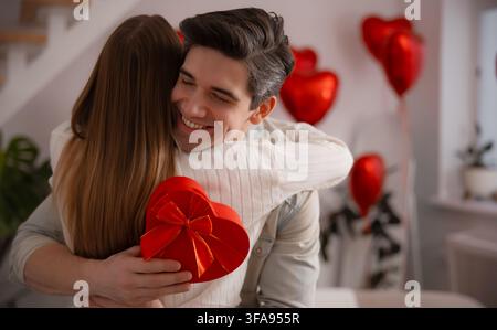 Glücklicher Mann umarmt seinen Partner, während er eine rote herzförmige Geschenkschachtel in einem Wohnzimmer hält, das mit schwebenden Herzballons dekoriert ist. Stockfoto