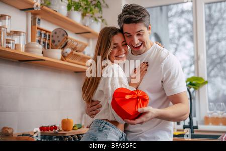 Fröhliches junges Paar umarmt sich in einer hellen Küche und lächelt auf eine rote herzförmige Geschenkschachtel hinab, die zwischen ihnen präsentiert wird. Stockfoto