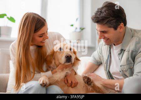 Ein liebevolles junges Paar lächelt seinen Golden Retriever an, während es den ruhigen Hund zusammen auf einem Sofa zu Hause kuschelt. Stockfoto