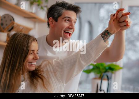 Nahaufnahme eines fröhlichen Paares, das seine verschränkten Hände erhebt, während es zusammen in einer hellen Küche lacht und tanzt. Stockfoto
