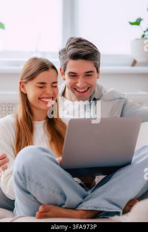 Fröhliches junges Paar lacht zusammen, während es einen Laptop anschaut und sich zu Hause auf dem Sofa entspannt Stockfoto